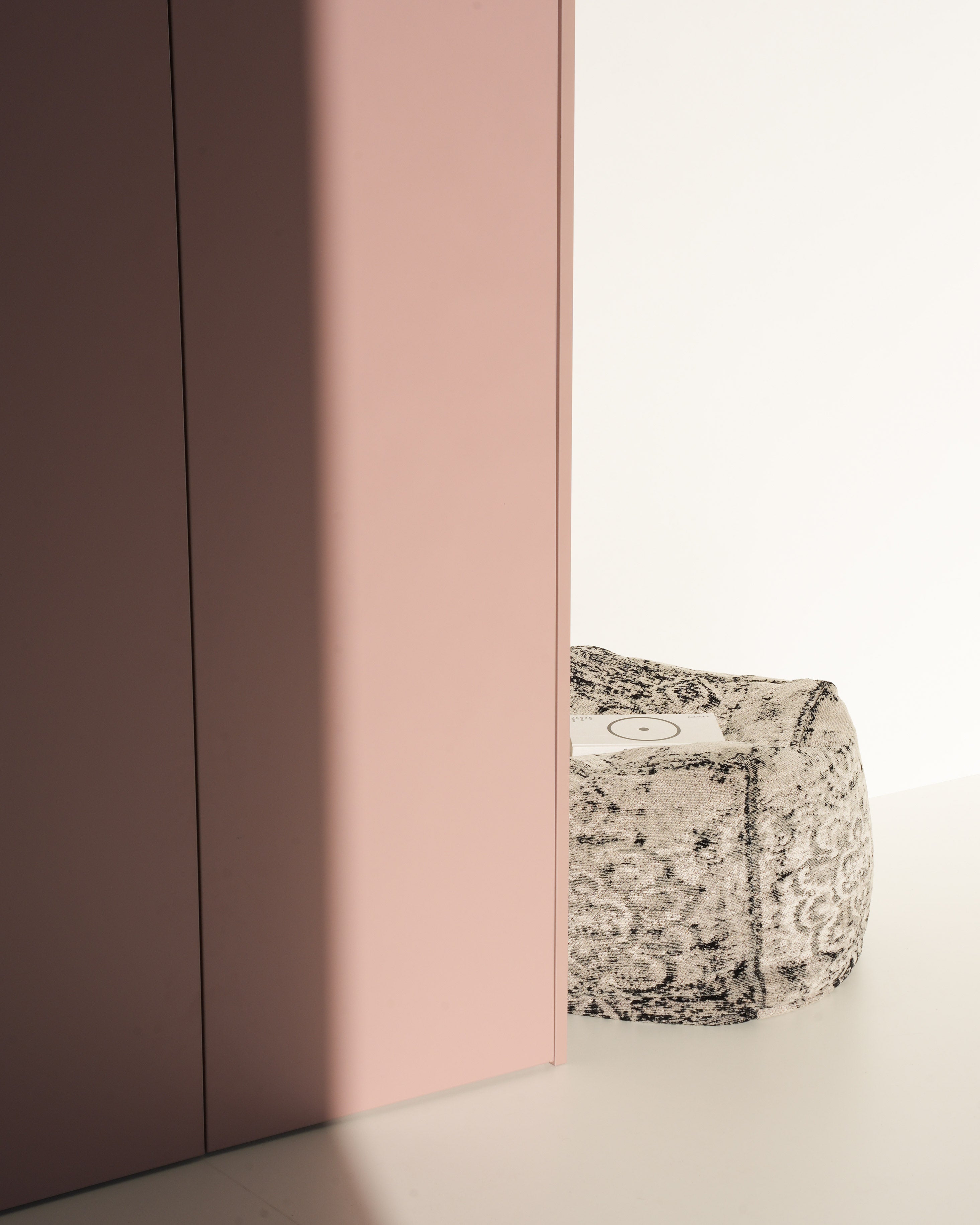 A blush pink cabinet stands next to a patterned grey and white floor pouf.