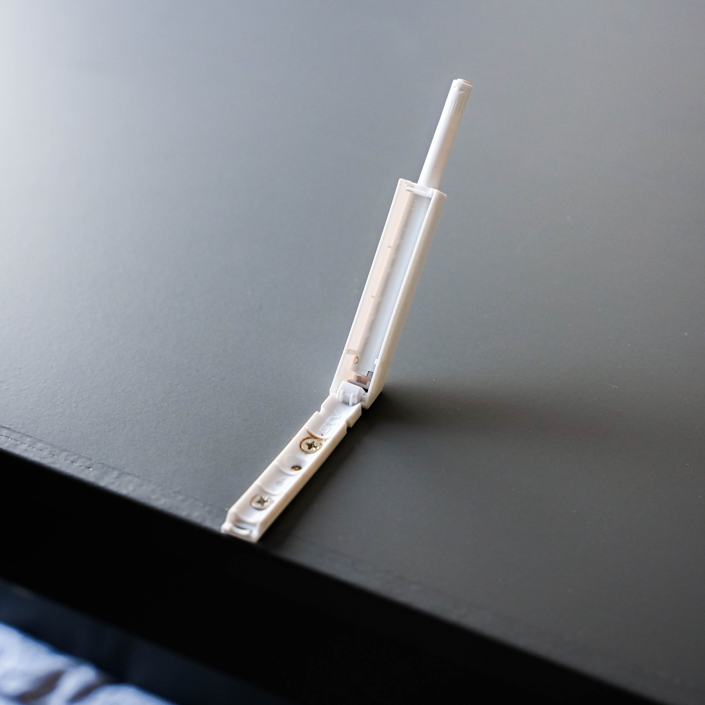 Ein weißer Tip-On-Drucktüröffner für PAX-Schränke, der an einer dunklen Oberfläche montiert ist.