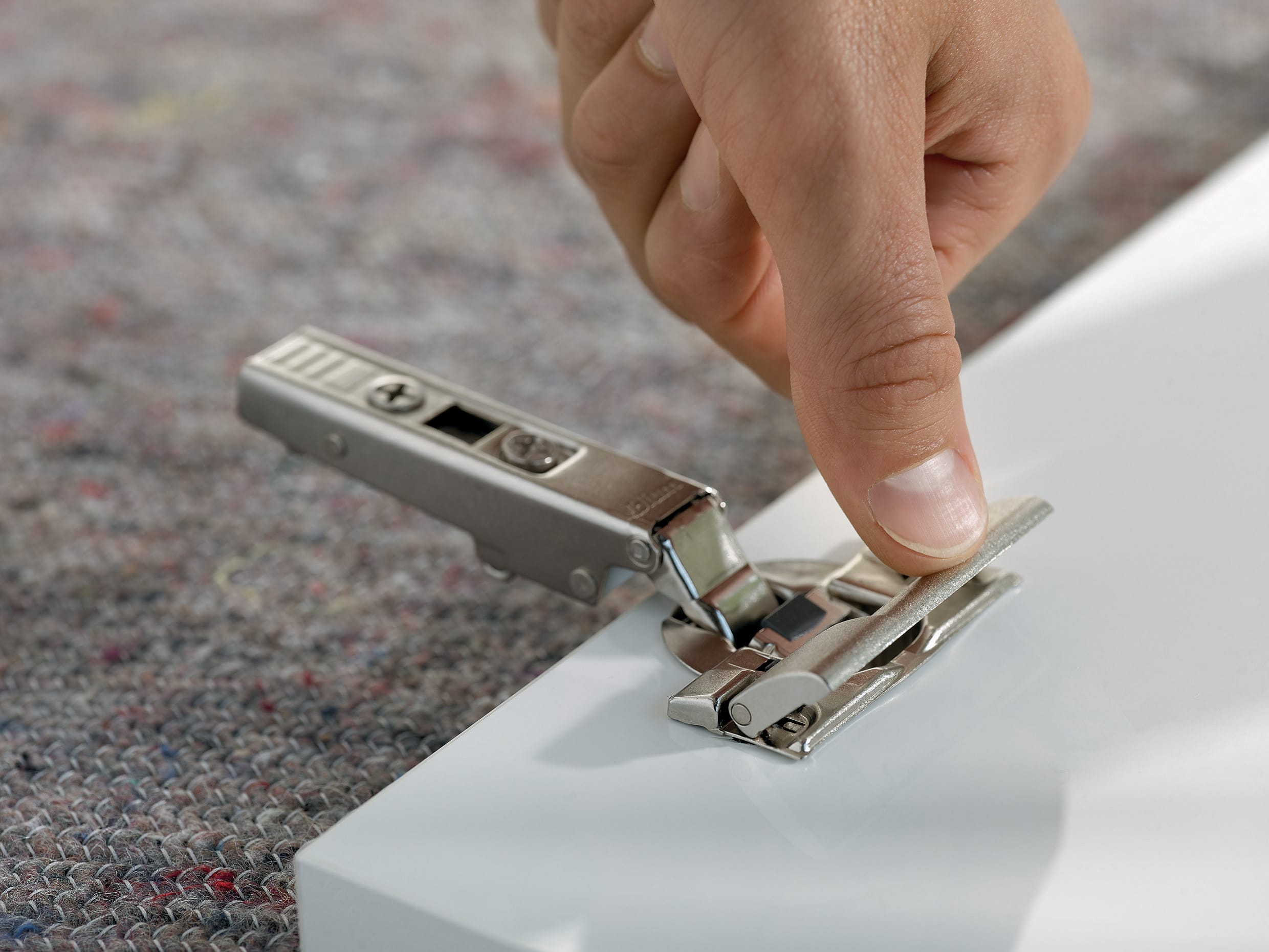 Eine Hand drückt ein silbernes Blum-Scharnier auf eine weiße Möbeltür.